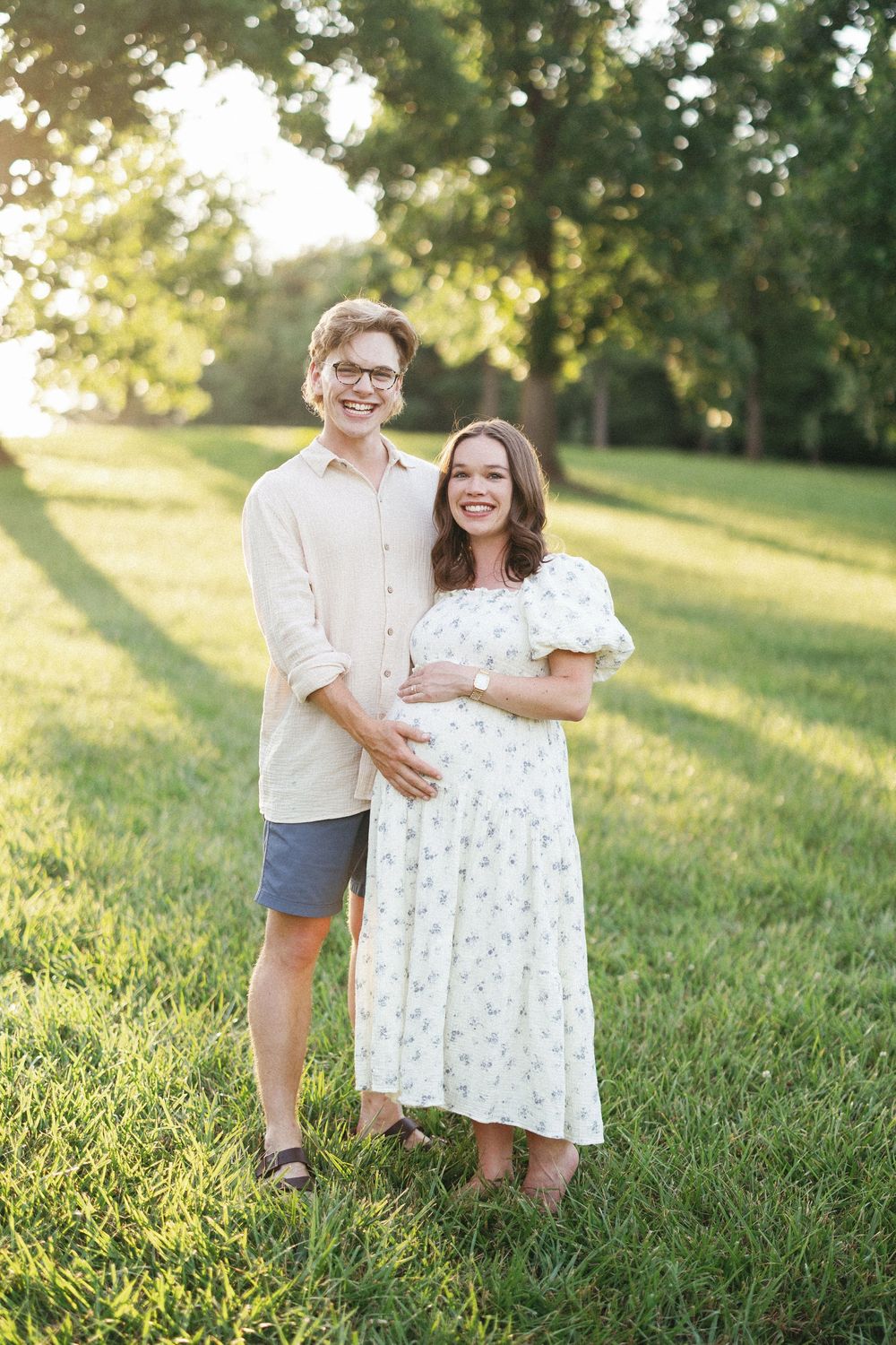 Smiling couple poses outdoors on green grass, woman in floral dress cradling baby bump, man in beige shirt, golden hour light.