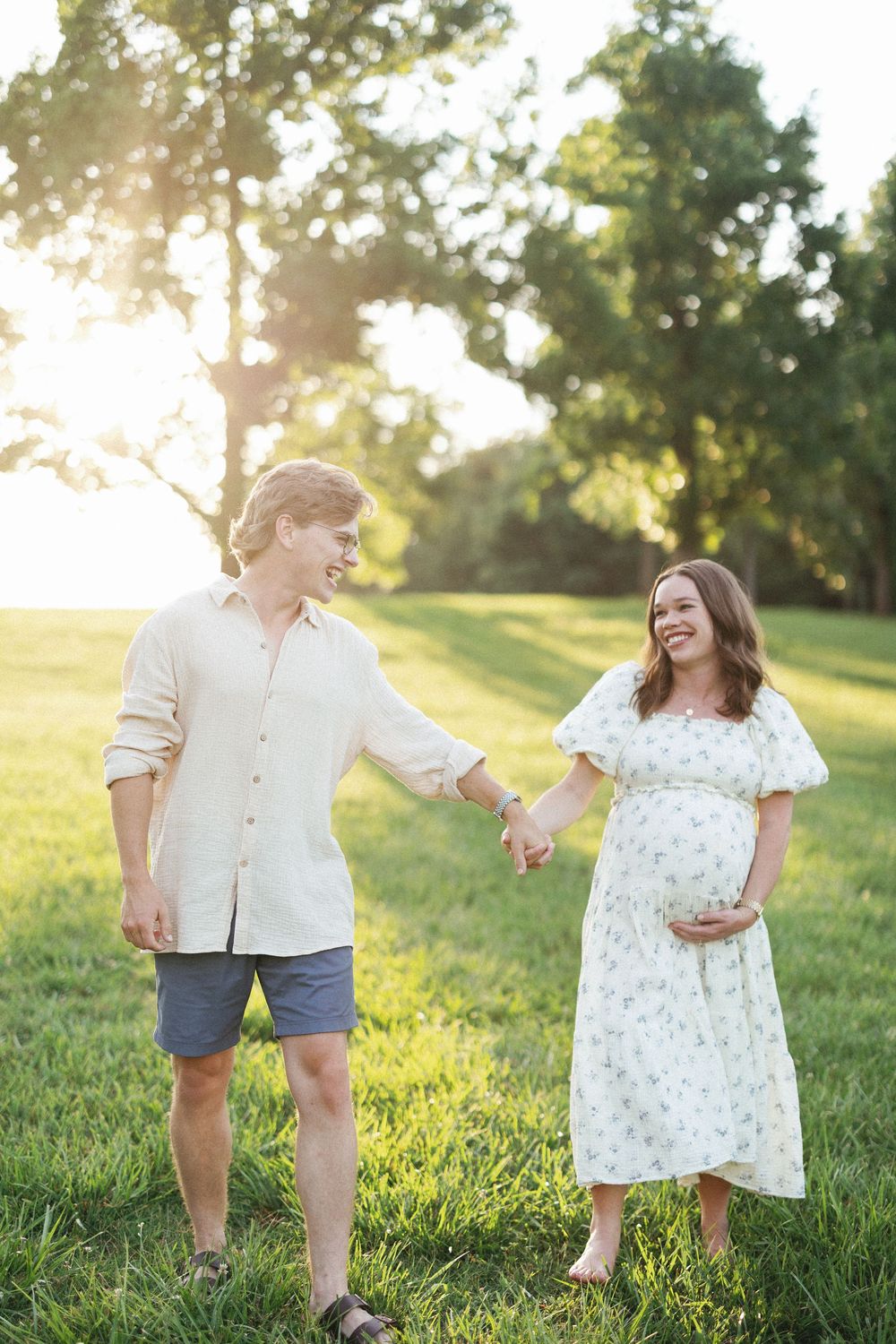 Smiling couple holding hands in a sunny park, pregnant woman in floral dress cradling her baby bump.