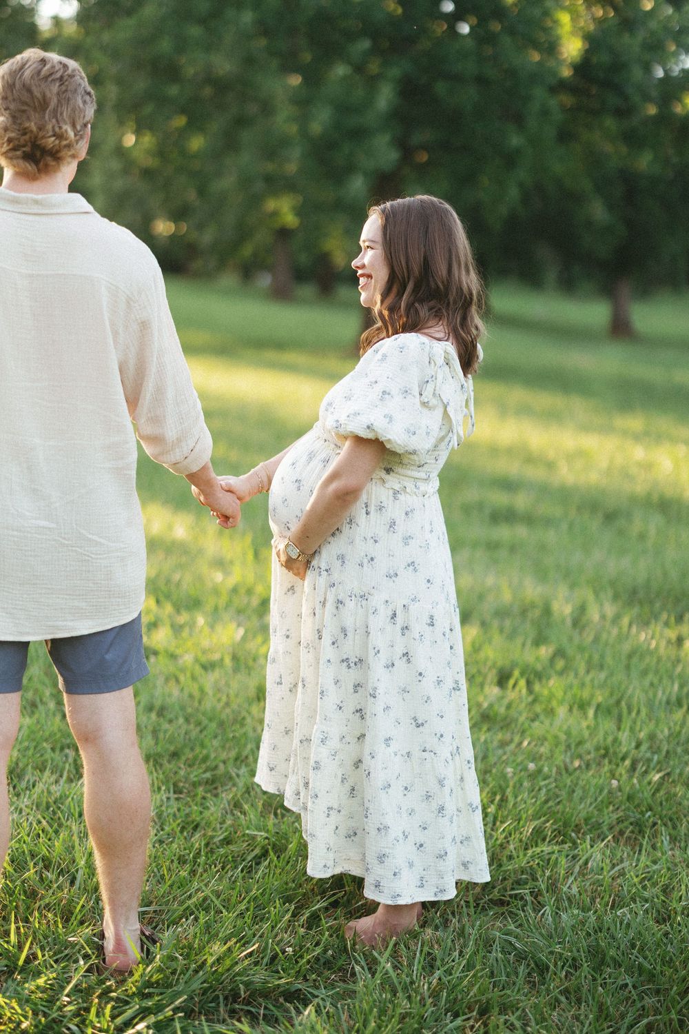 Pregnant woman in floral dress holds hands with partner in sunny green field, smiling joyfully during maternity photoshoot.