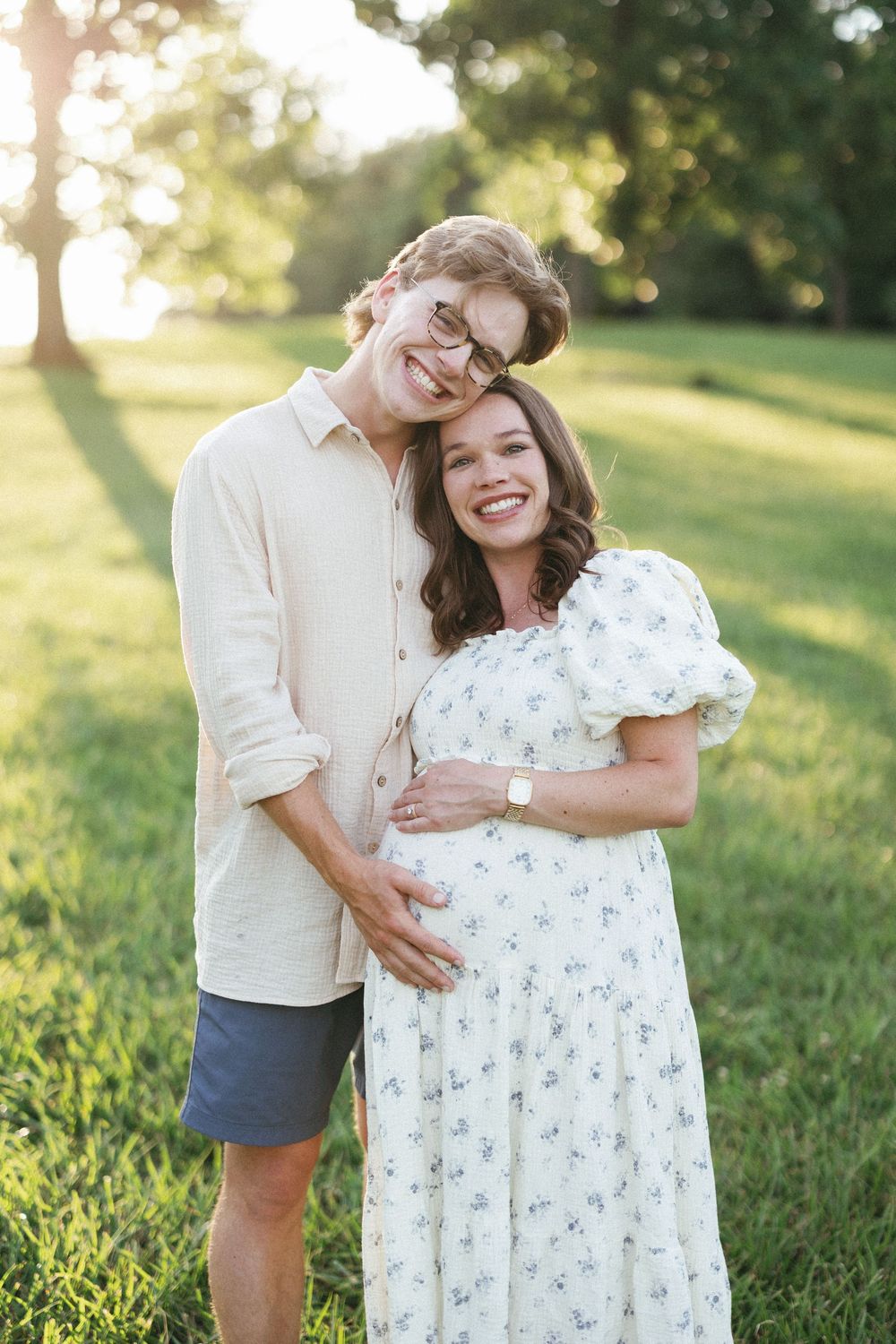 Happy couple smiling outdoors in golden hour light, pregnant woman in floral dress cradled by partner in green park setting.