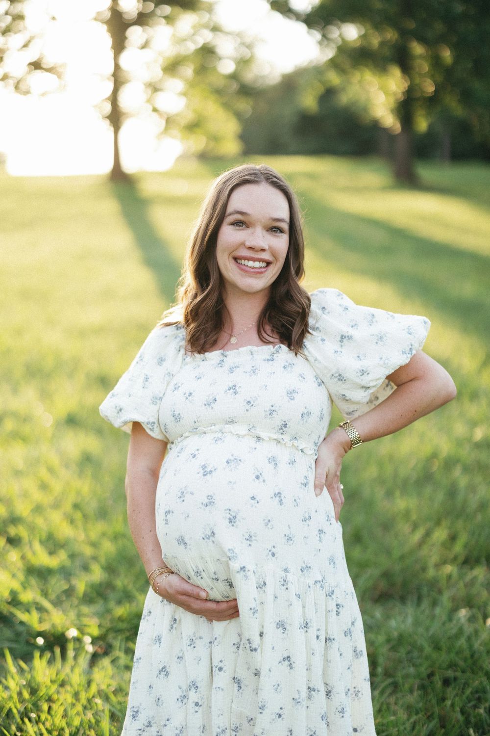 Smiling pregnant woman in white floral dress cradling baby bump outdoors in sunlit green park.