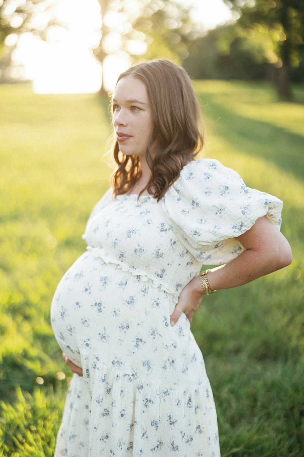 Pregnant woman in floral white dress holding baby bump in sunlit green field during golden hour.