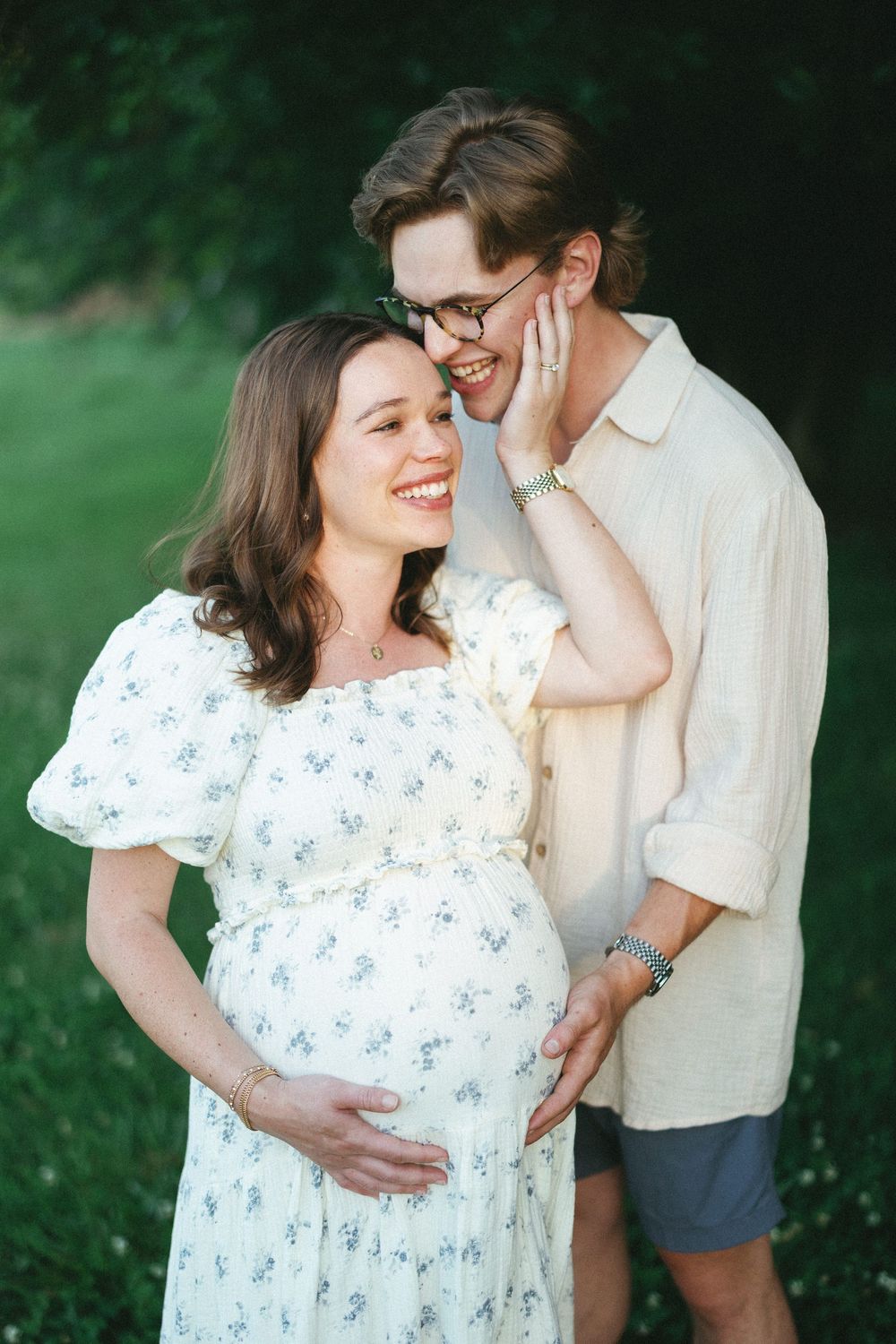 Smiling pregnant woman in floral dress with partner tenderly touching her face outdoors.