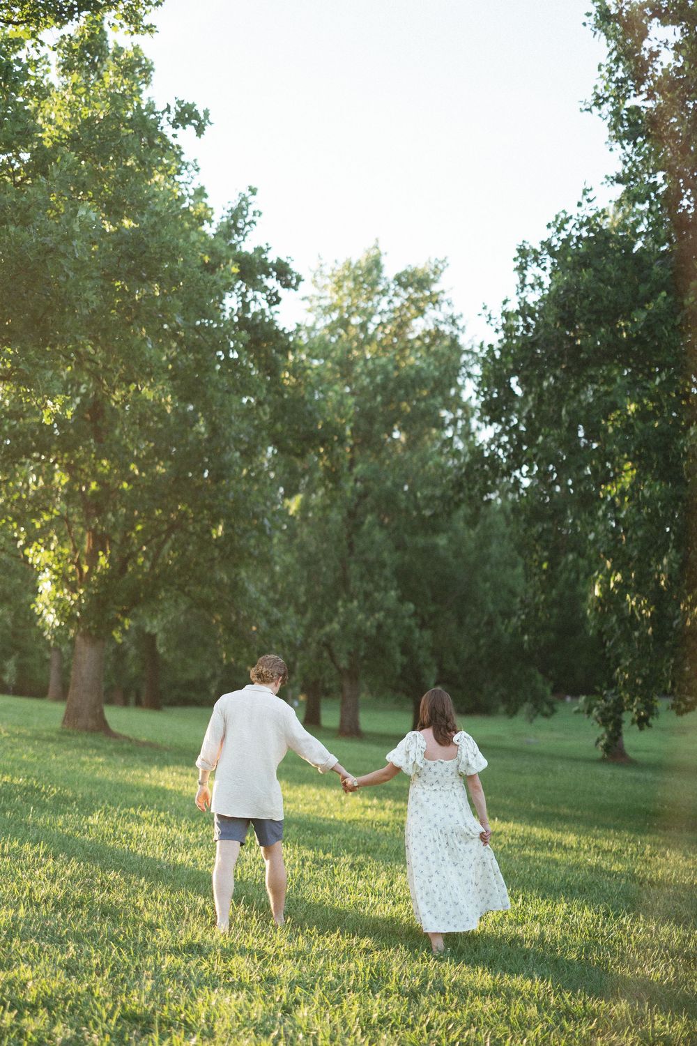 Couple holding hands walking through a lush green park on a sunny day, woman in floral dress and man in white shirt.
