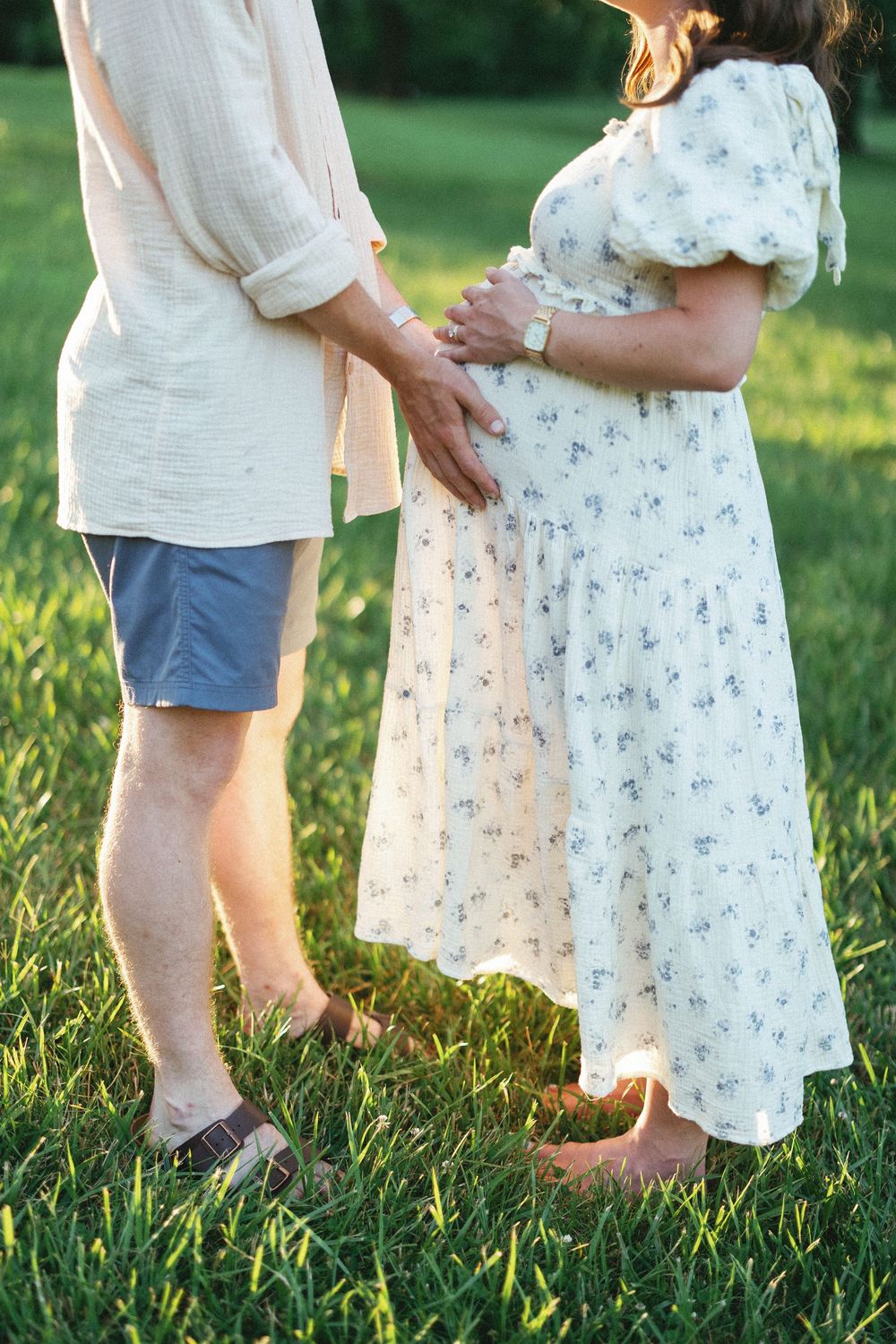 Close-up of couple holding pregnant belly together on sunny green lawn.