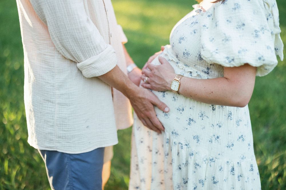 Couple tenderly holding pregnant belly outdoors in green field, woman in floral dress with gold watch.