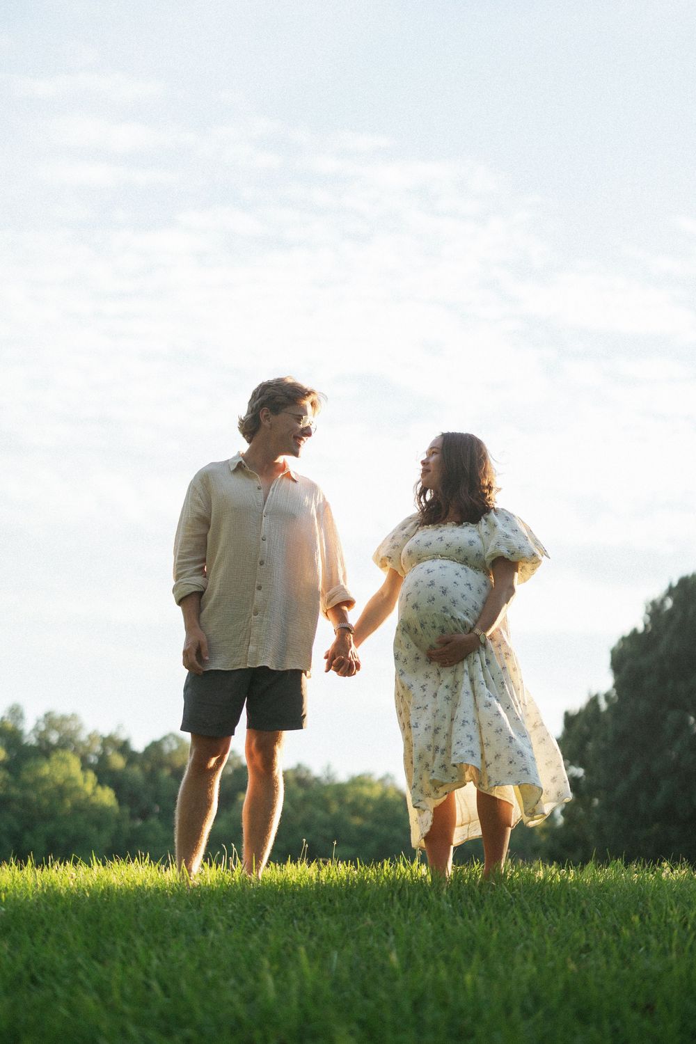 A pregnant couple holds hands on a sunny green lawn, smiling at each other, with trees and blue sky behind them.