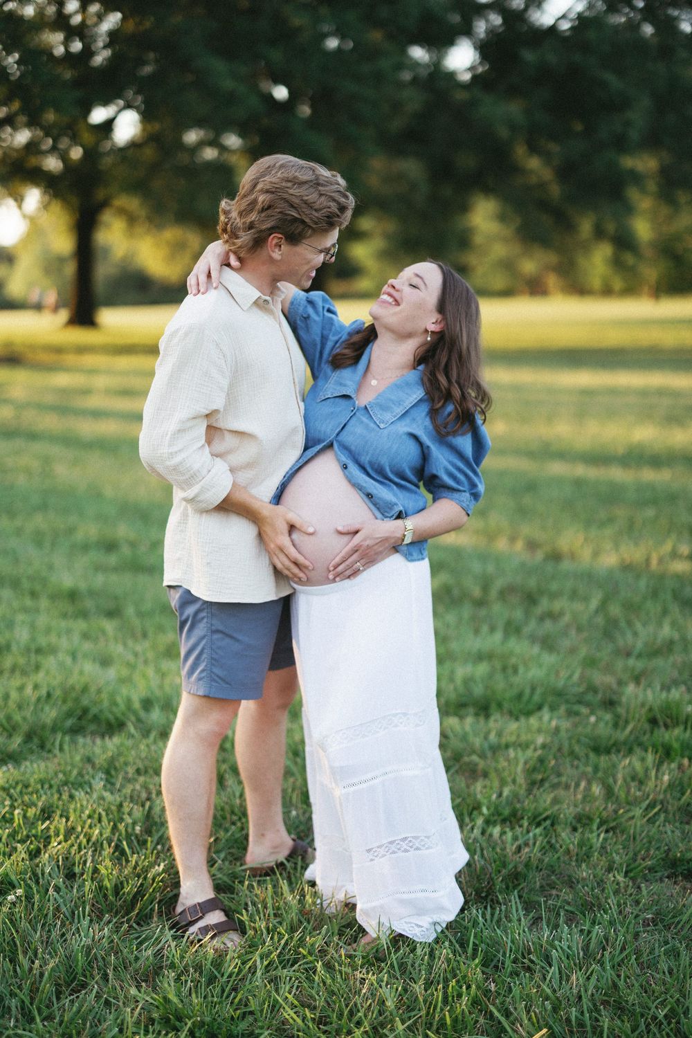 Couple cradling pregnant belly outdoors in a sunny green park, woman in denim shirt and white skirt smiling up at partner.