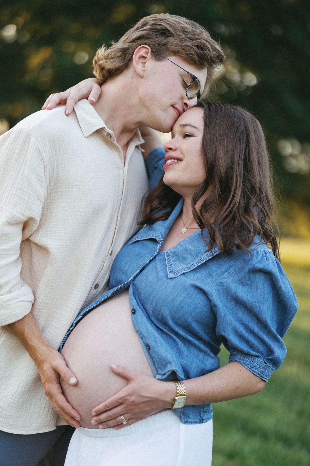 Couple sharing tender moment outdoors, pregnant woman in denim shirt cradling baby bump while partner kisses her forehead.
