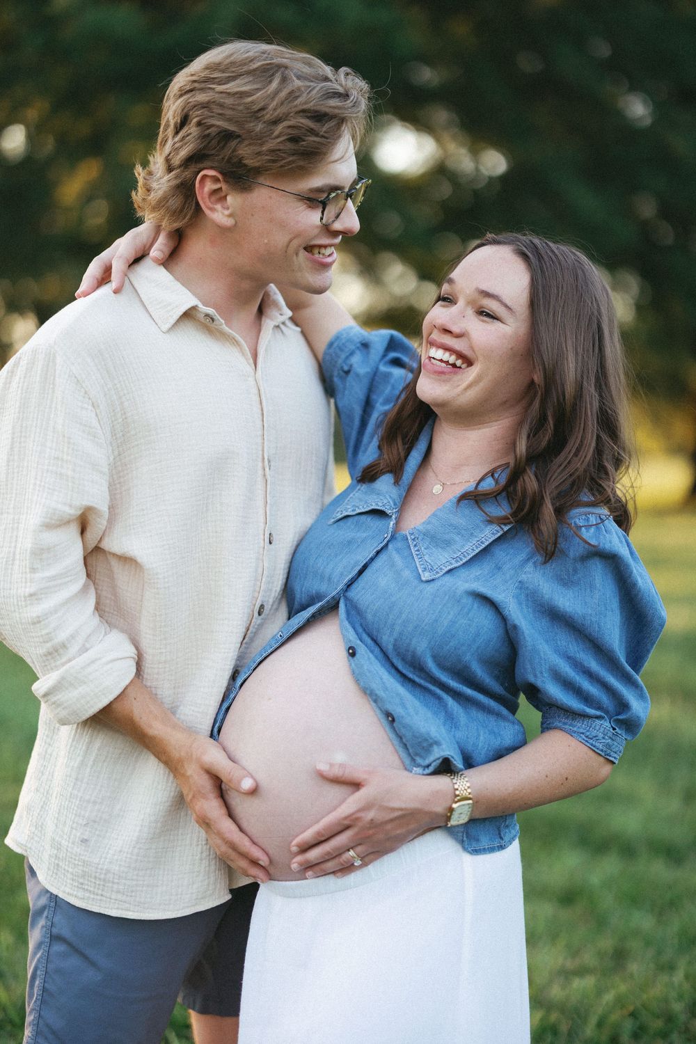 Happy pregnant couple embracing outdoors, woman in denim shirt laughing while cradling baby bump, man in glasses smiling beside her.