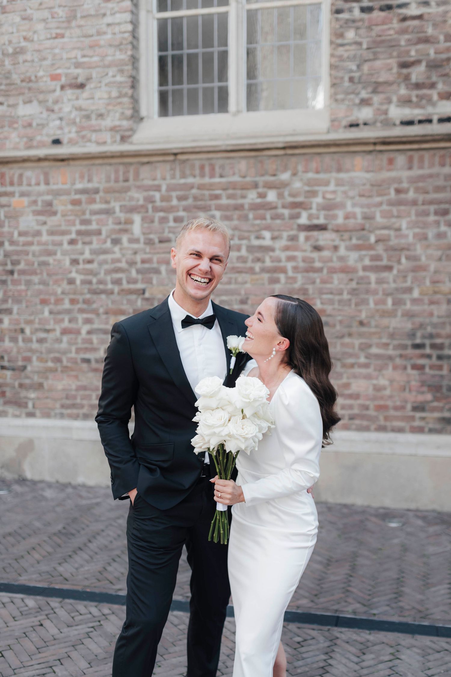 Bride and groom laughing together outdoors, she holds white peonies bouquet, he wears black tuxedo, brick building backdrop.