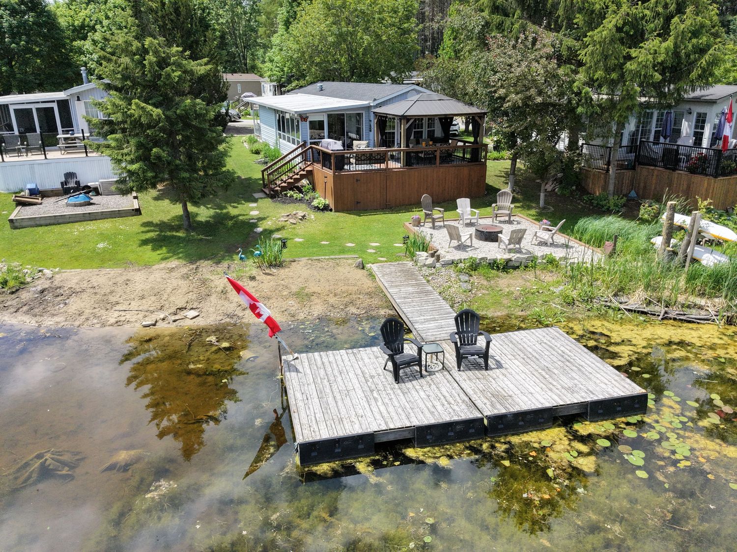 Aerial view of a lakeside cottage with a wooden dock, Adirondack chairs, fire pit area, and lush green trees.