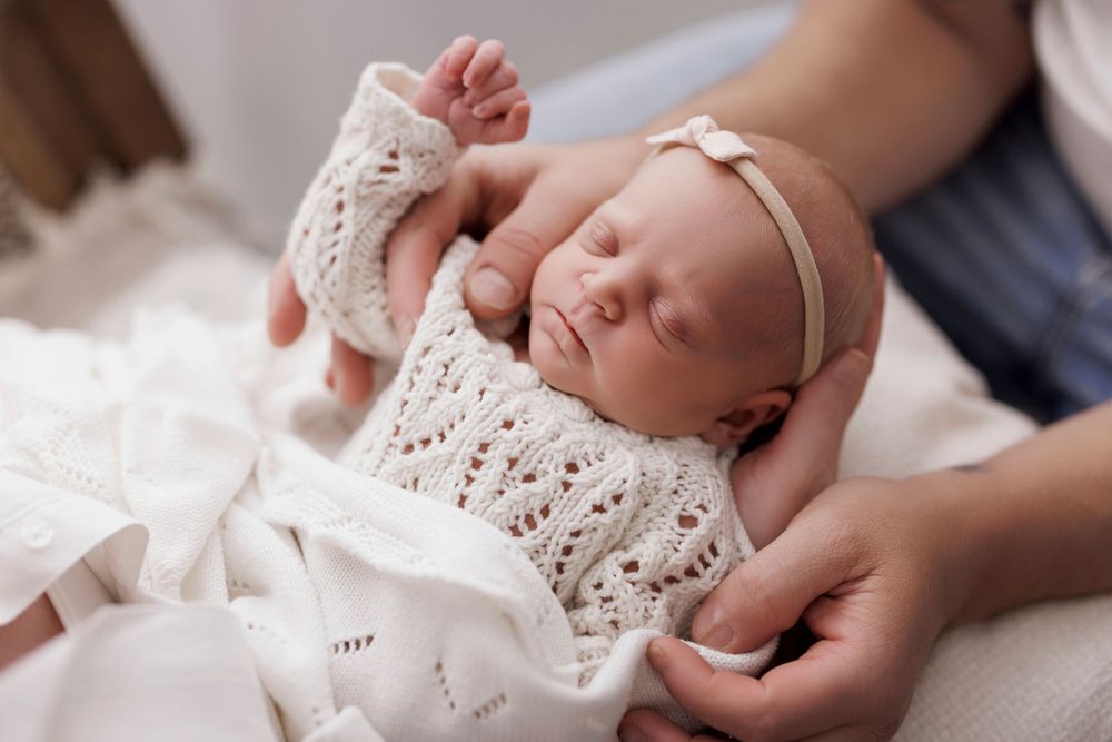 Newborn baby sleeping peacefully in white knit outfit and bow headband, cradled gently in adult hands.