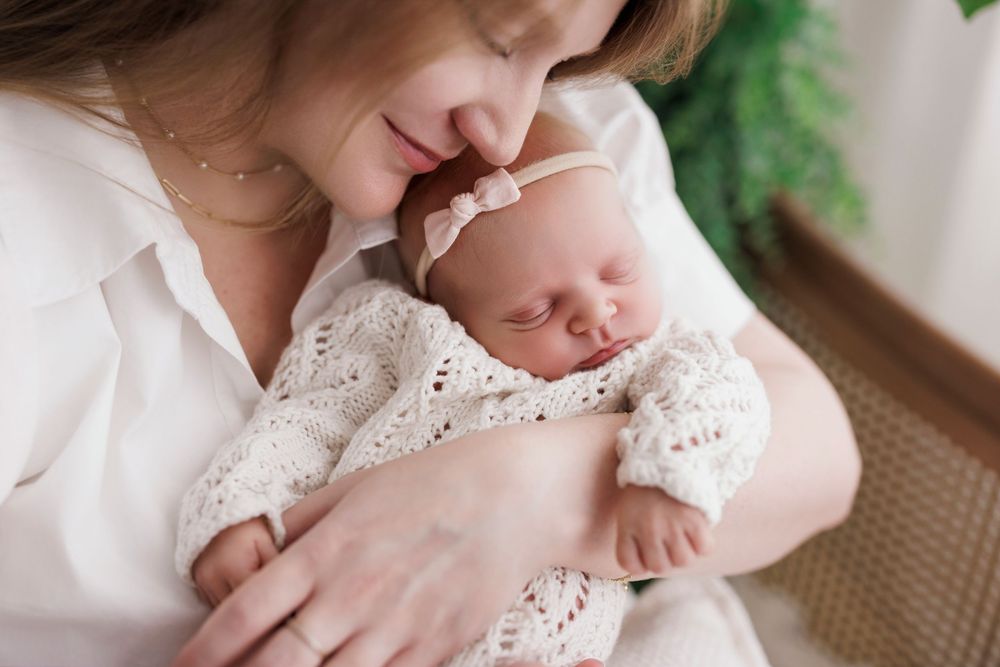 Mother tenderly holds sleeping newborn baby dressed in white lace outfit with pink bow headband.