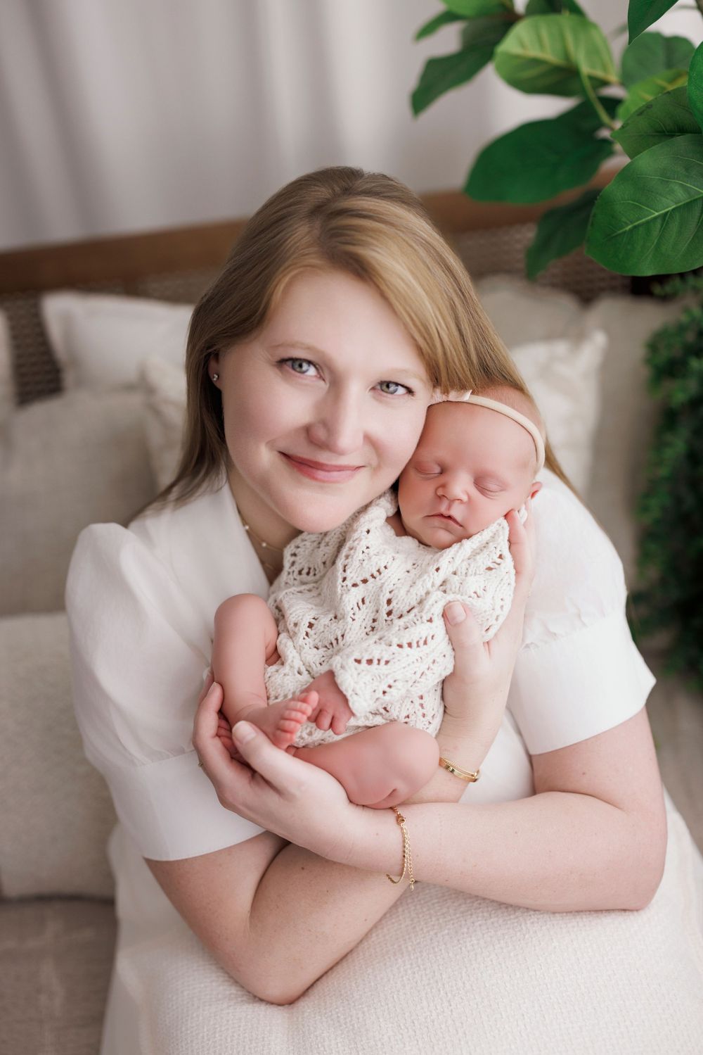 Smiling mother in white dress tenderly holds sleeping newborn baby wrapped in cream knit blanket.