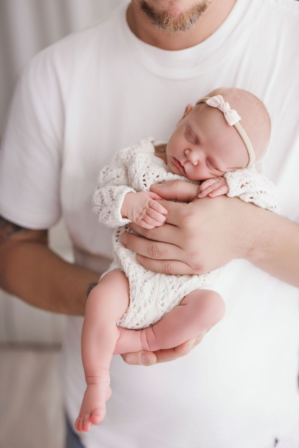 A father in a white t-shirt gently cradles his sleeping newborn baby girl dressed in a white lace outfit with a bow headband.