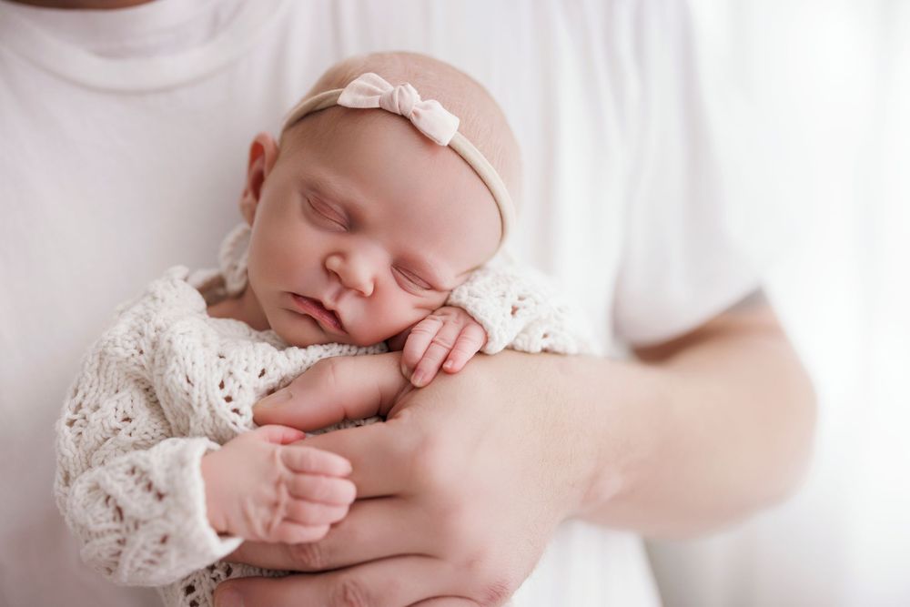 Newborn baby girl sleeping peacefully in parent's arms, wearing white lace outfit and bow headband.