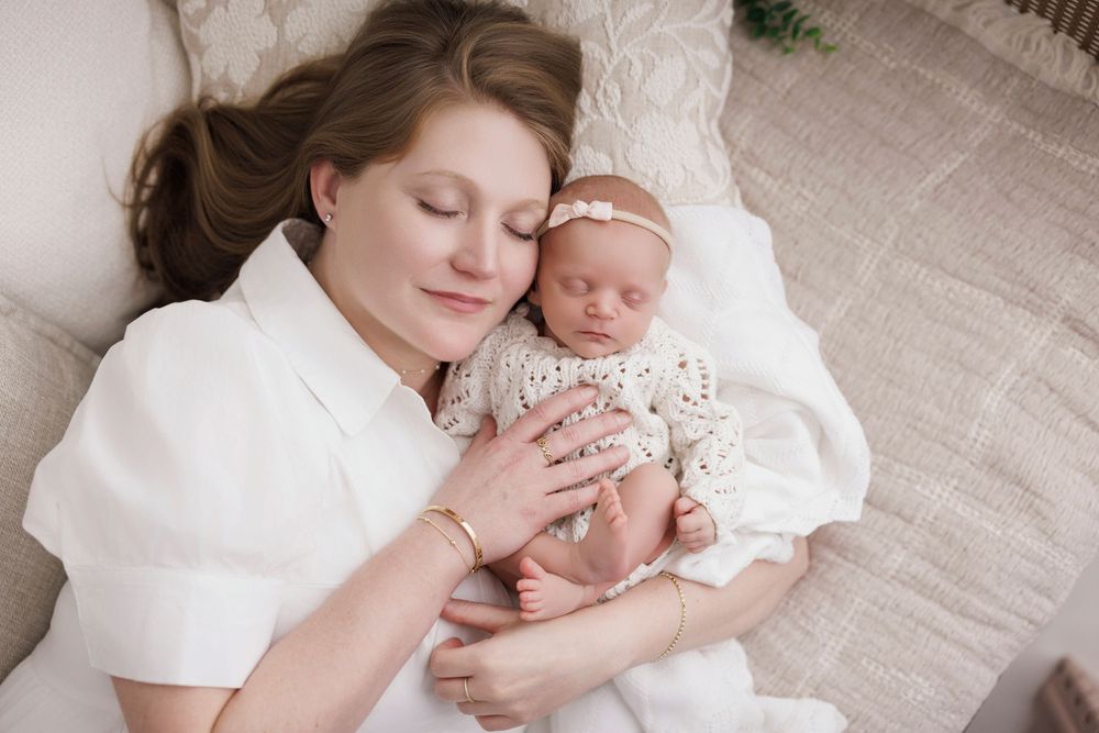 Mother lying down with eyes closed, tenderly holding her sleeping newborn baby dressed in a white crochet outfit.