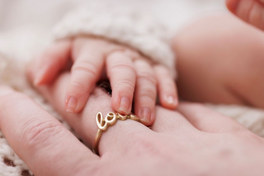 A newborn baby's tiny hand resting on a mother's finger wearing a delicate gold 'love' ring.