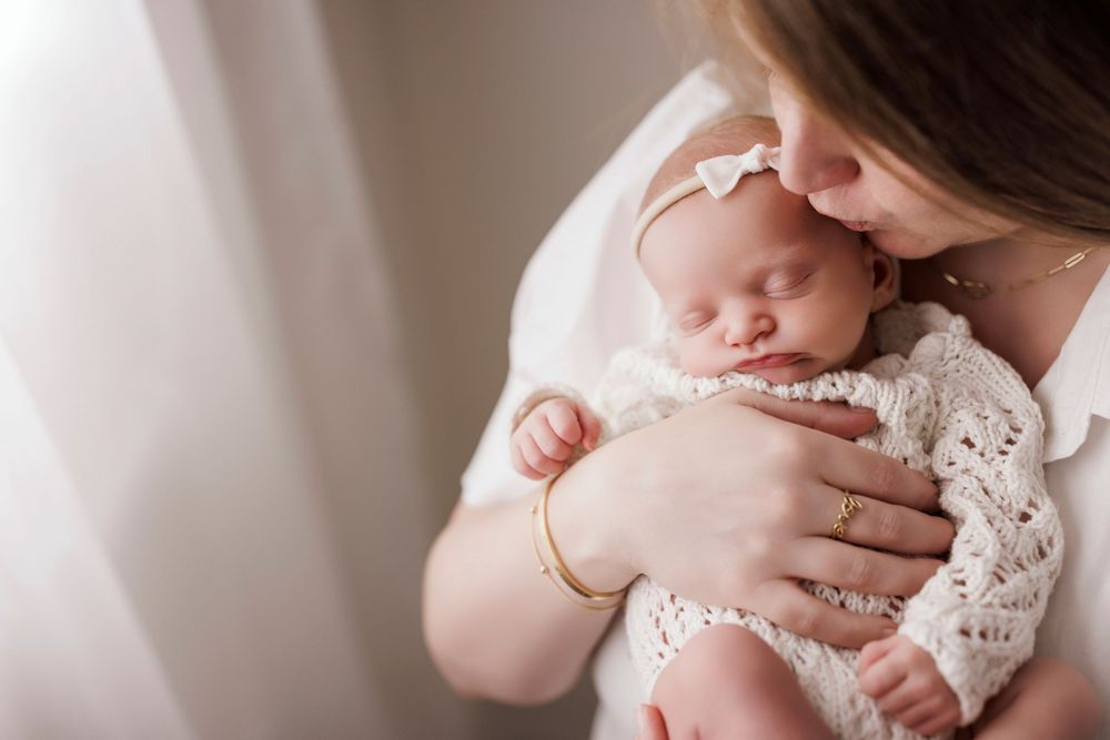 Mother tenderly kissing her sleeping newborn baby wrapped in white lace, soft natural light photography.