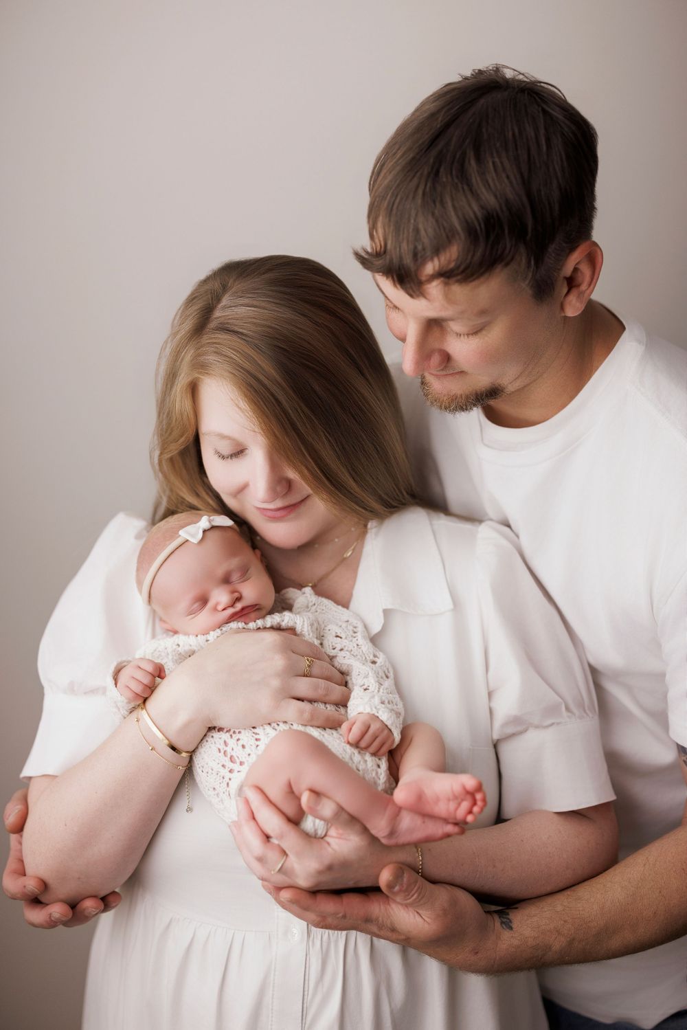 Happy couple in white outfits tenderly holds their sleeping newborn baby dressed in white lace outfit.