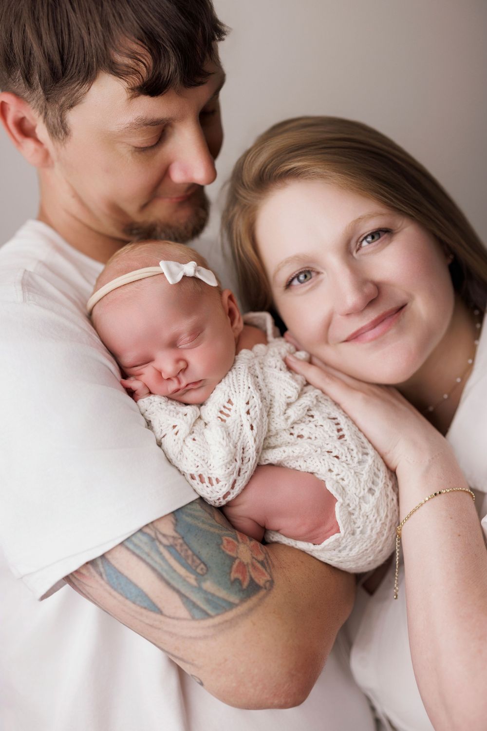 Happy parents in white shirts lovingly cradle their sleeping newborn wrapped in cream knit swaddle.