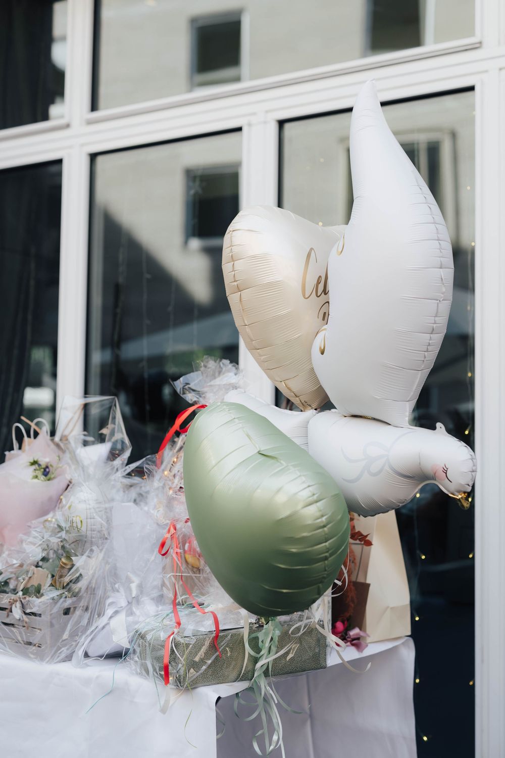 Elegant celebration balloons in white, gold, and sage green displayed with floral arrangements near a glass building exterior.