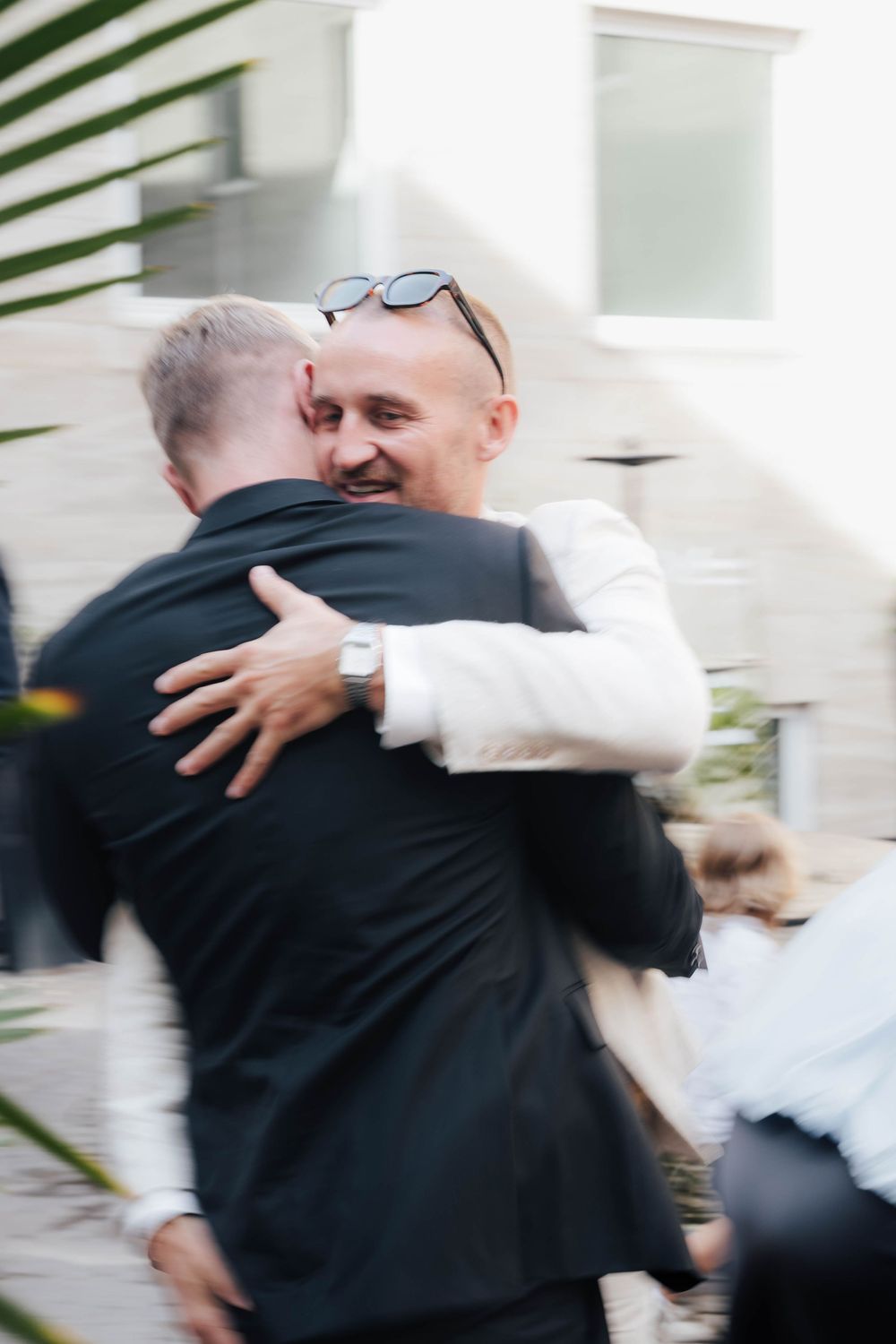 Two men in formal suits share a warm embrace outdoors, one smiling with sunglasses on his head.