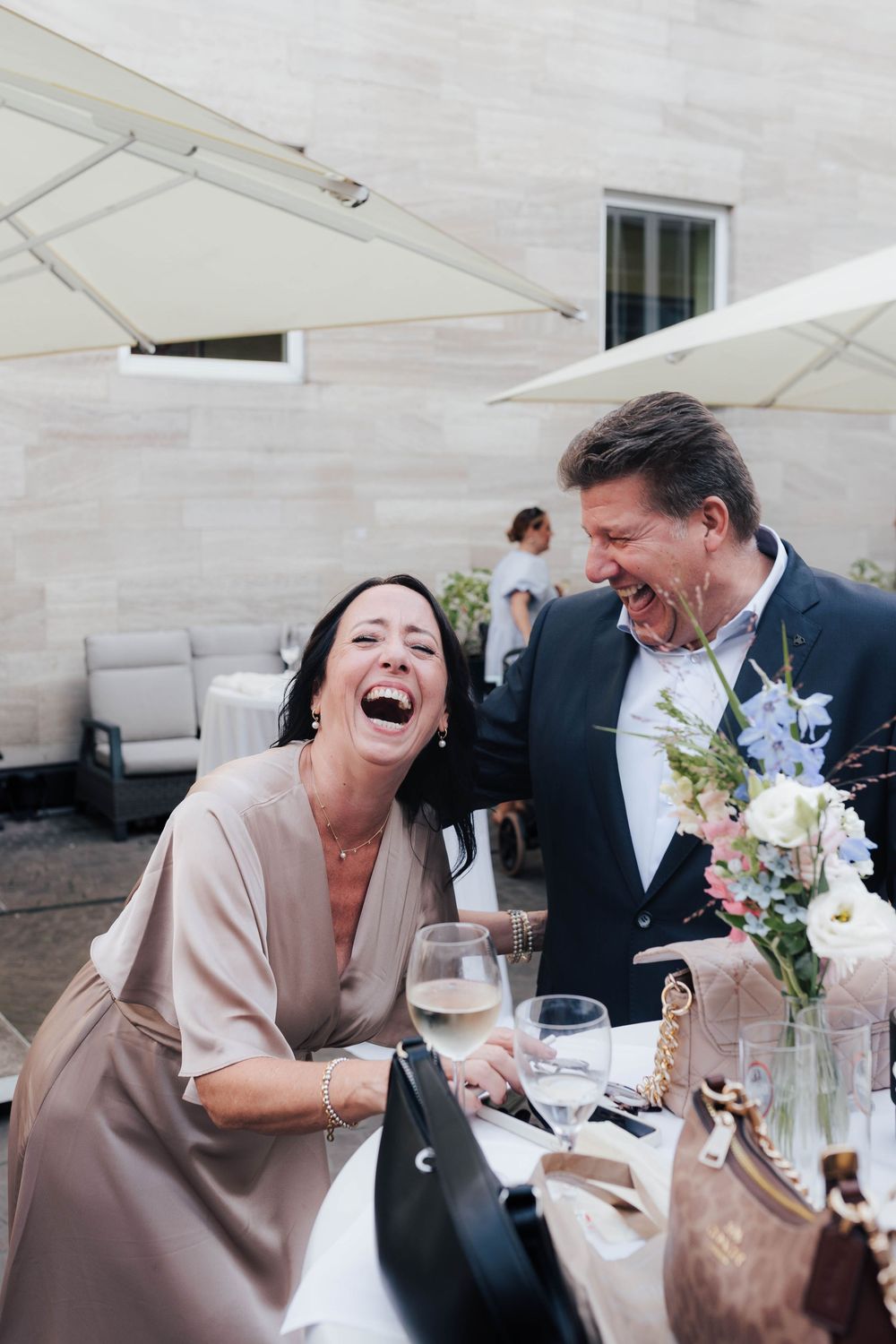 Couple laughing joyfully at outdoor wedding reception table with floral centerpiece.