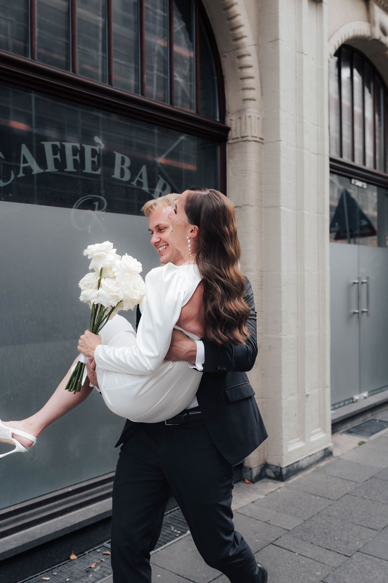 Groom carries bride holding white flowers outside a café bar on a city street in elegant black and white wedding attire.