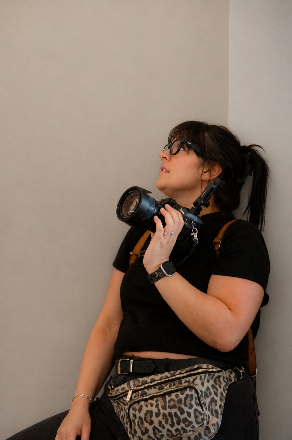 Woman in black top holding DSLR camera, wearing sunglasses and leopard print fanny pack, leaning against wall.