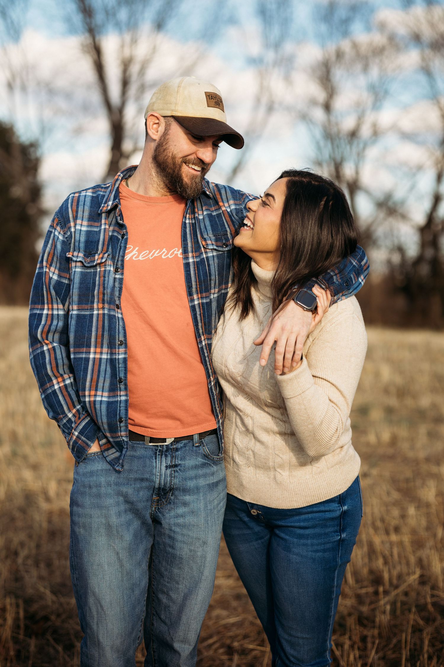 A smiling couple in casual flannel and jeans embracing outdoors in a field during autumn.