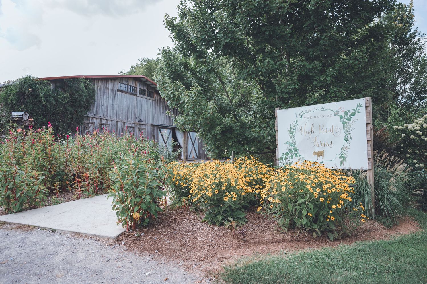 Rustic wooden barn surrounded by lush trees and vibrant yellow wildflowers with a decorative garden sign in front.
