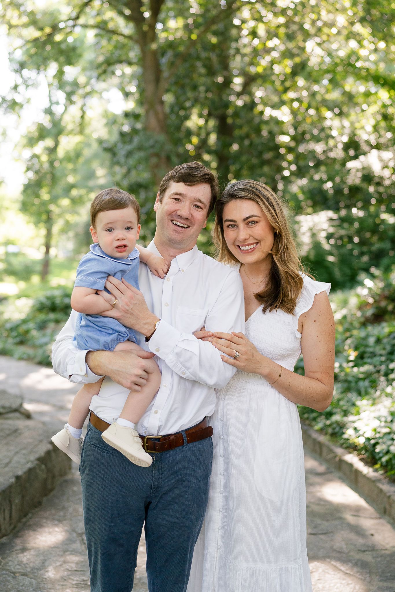 Happy family portrait outdoors with father holding baby boy and mother in white dress smiling in lush green park.