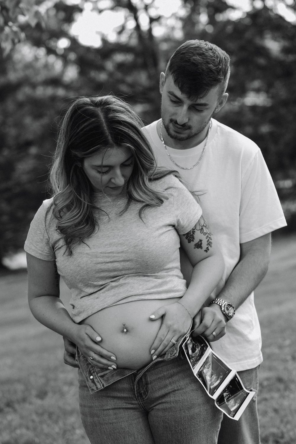 Pregnant mother and father holding and looking at baby bump in a grassy sunny field while holding an ultrasound