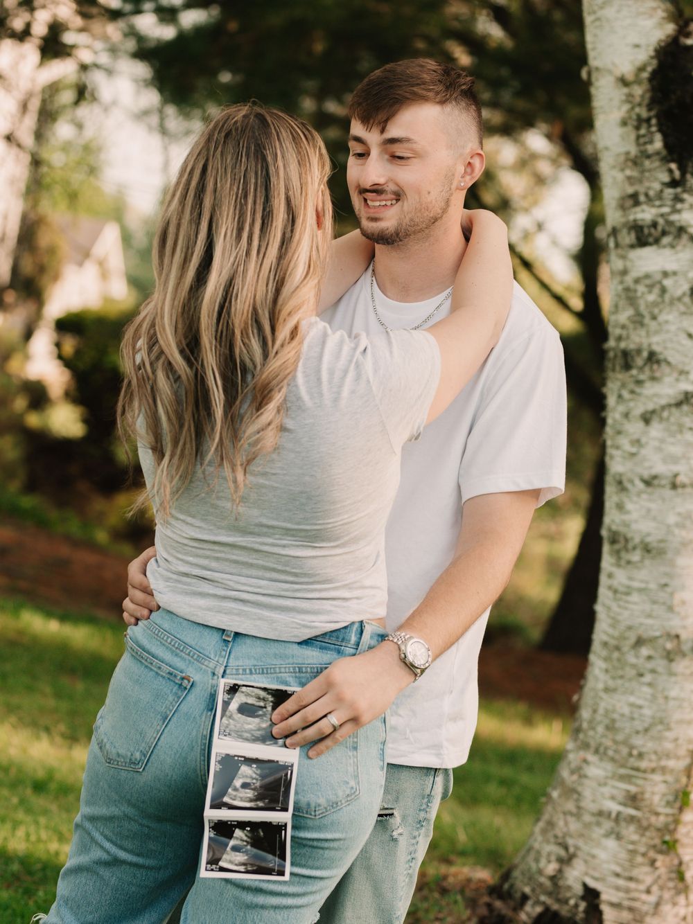 Pregnant mother and father gazing at each other in a grassy field near a birch tree while holding an ultrasound toward the camera