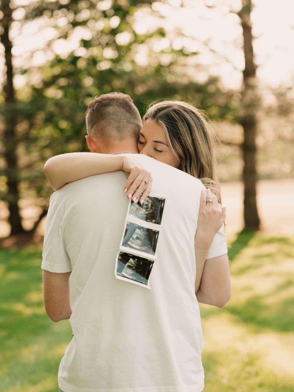 Pregnant mother and father embrace in a grassy sunny field while holding an ultrasound toward the camera