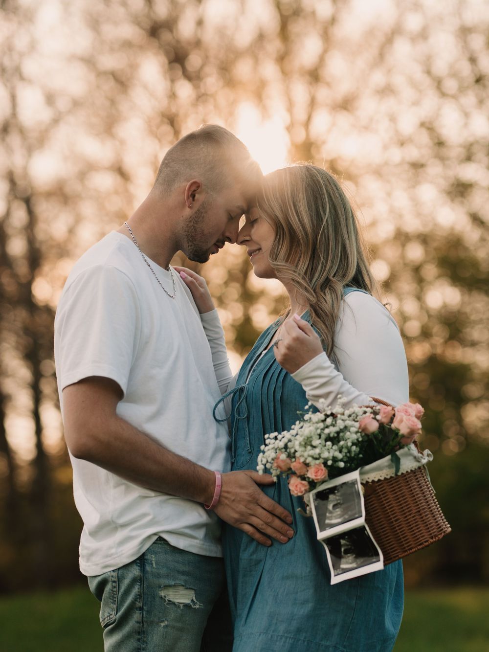 Pregnant woman and father standing nose to nose in a golden, sunny forest, holding a basket of pink flowers with an ultrasound on the side