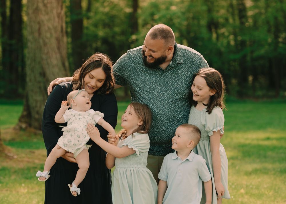 Family of 6 six standing in a sun-lit grassy field making a baby girl laugh