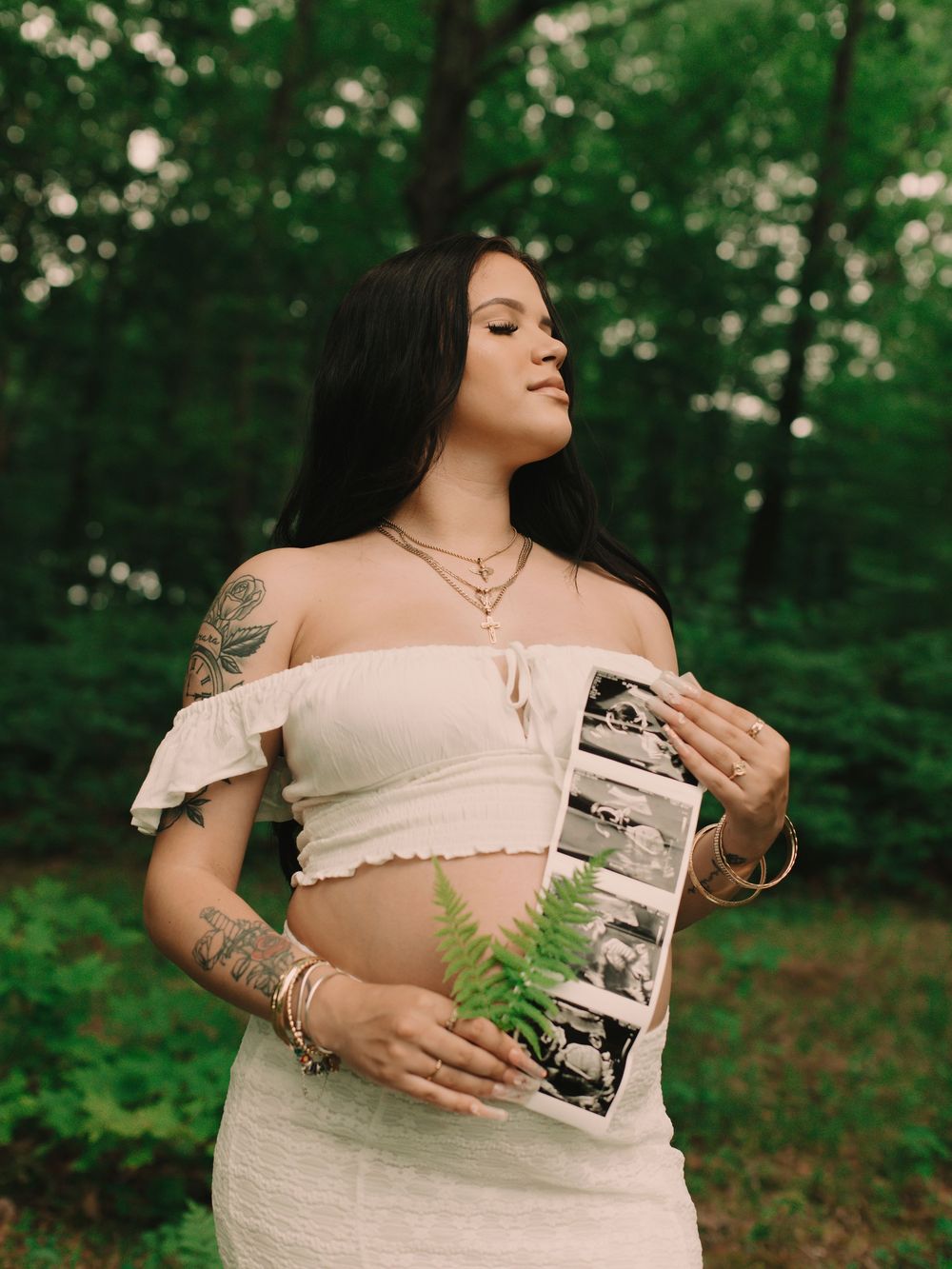 Pregnant mother holding an ultrasound with ferns in front of her baby bump outdoors in the forest