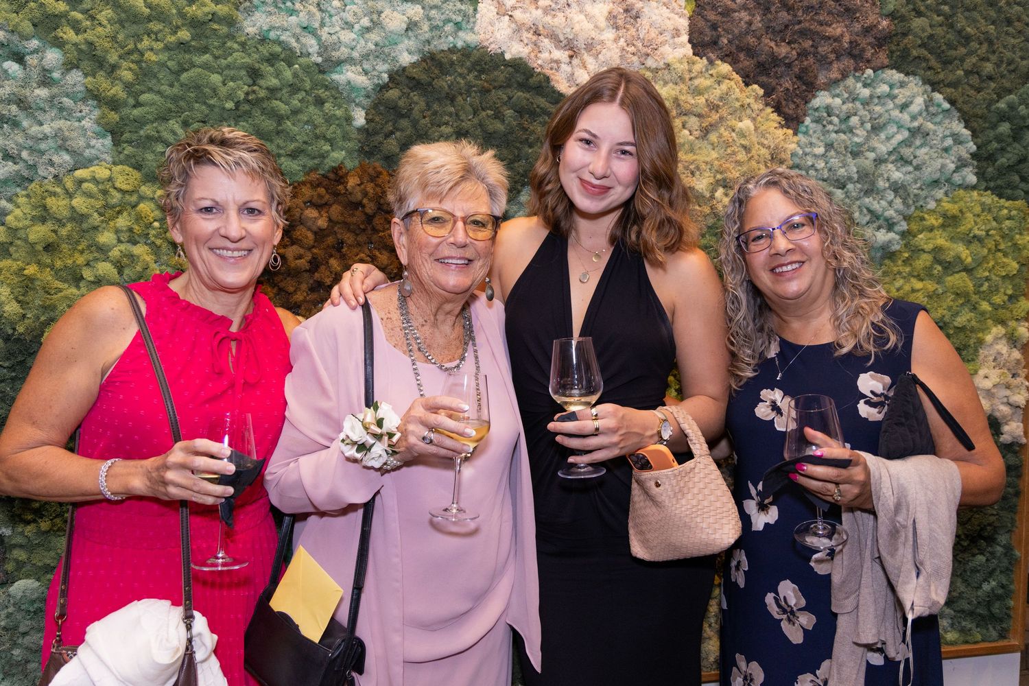Four women smiling and holding wine glasses at a wedding reception with a green moss wall backdrop in the Alcove Cincinnati wedding venue.