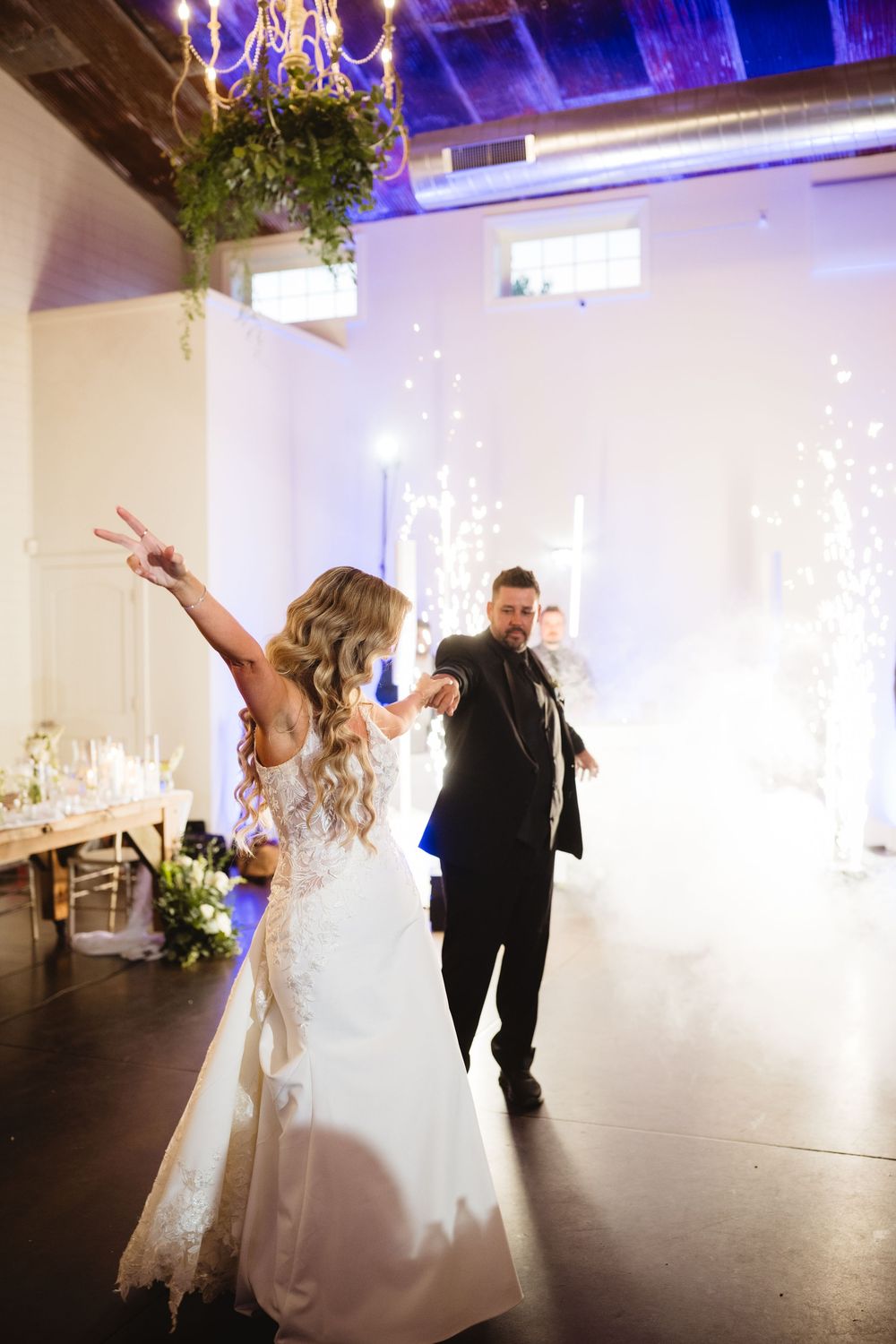 Bride and groom make grand entrance at wedding reception with sparklers and fog effects in elegant white venue.