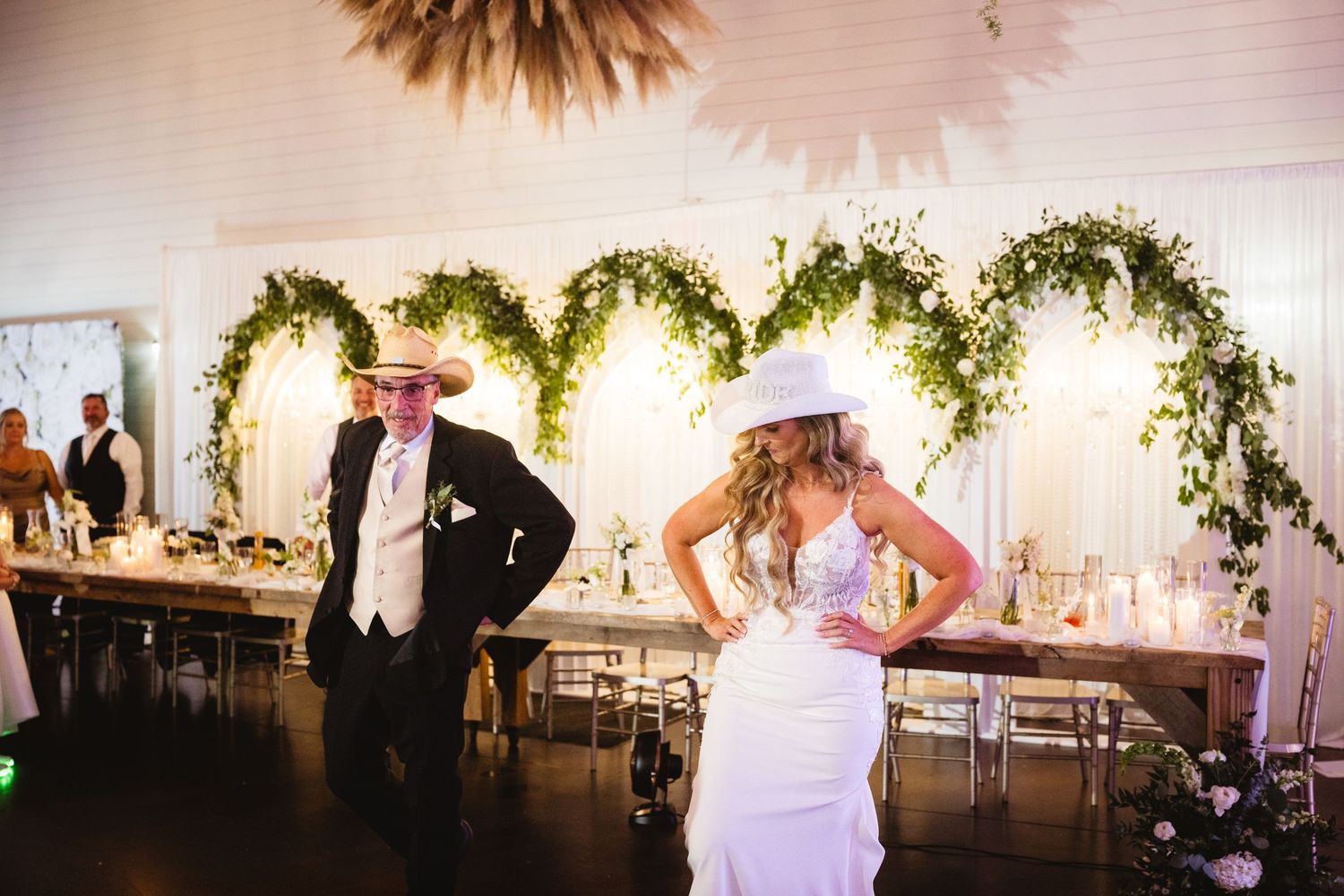 Bride and groom dancing at rustic wedding reception with heart-shaped greenery backdrop and elegant table settings.