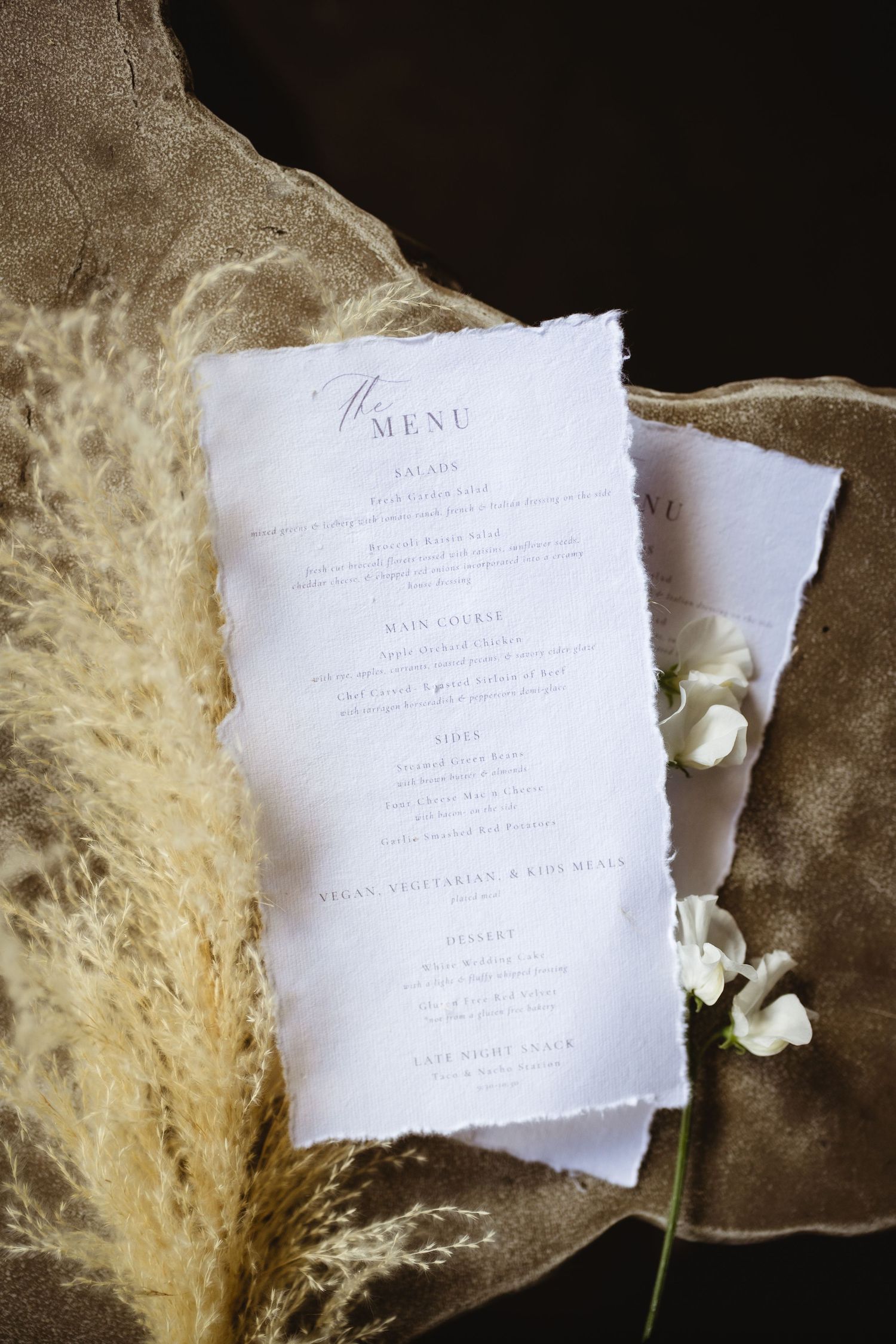 Elegant wedding dinner menu card with deckled edges displayed on velvet with pampas grass and white flowers.