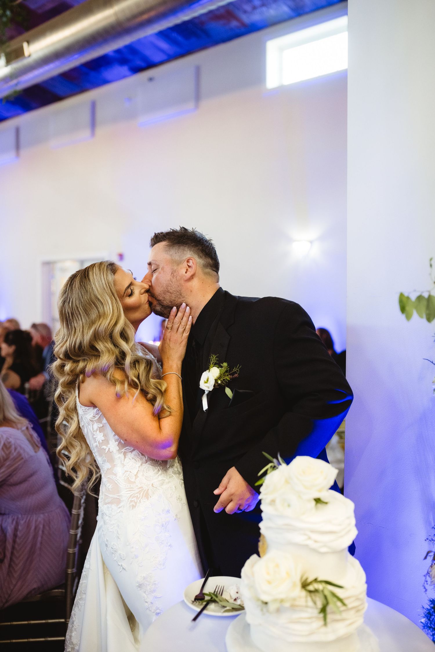 Bride and groom share a kiss beside their white wedding cake at their elegant reception venue.