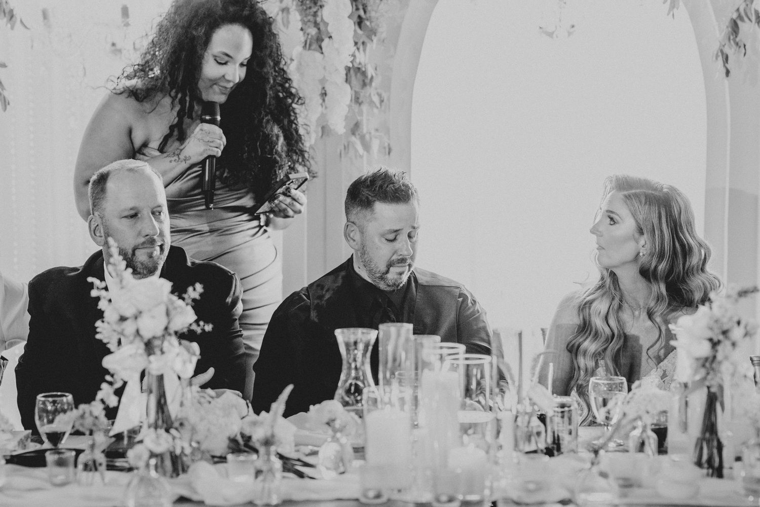 Wedding guests laughing together at reception table with floral arrangements in black and white photo.