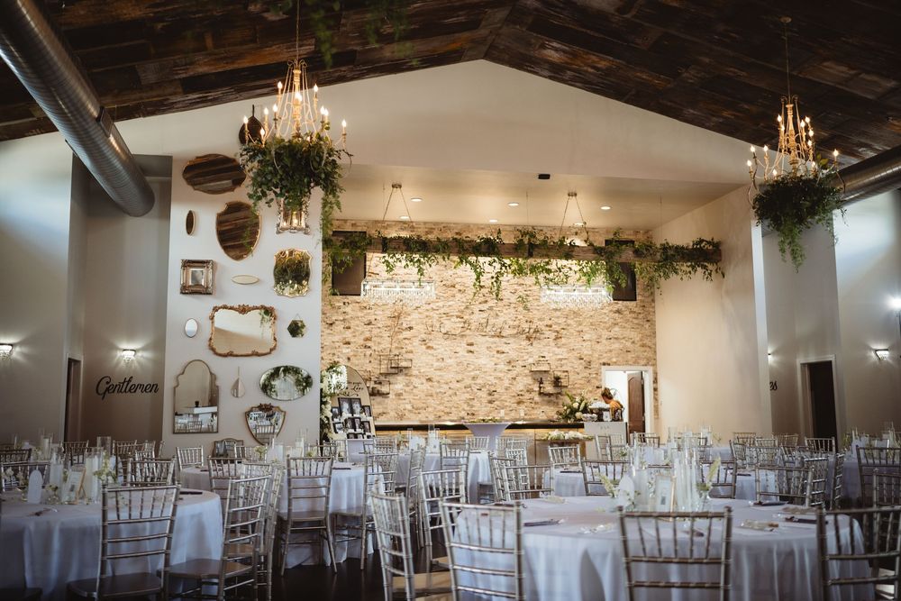 Elegant wedding reception hall with white chiavari chairs, round tables, exposed brick wall, and lush greenery chandeliers.