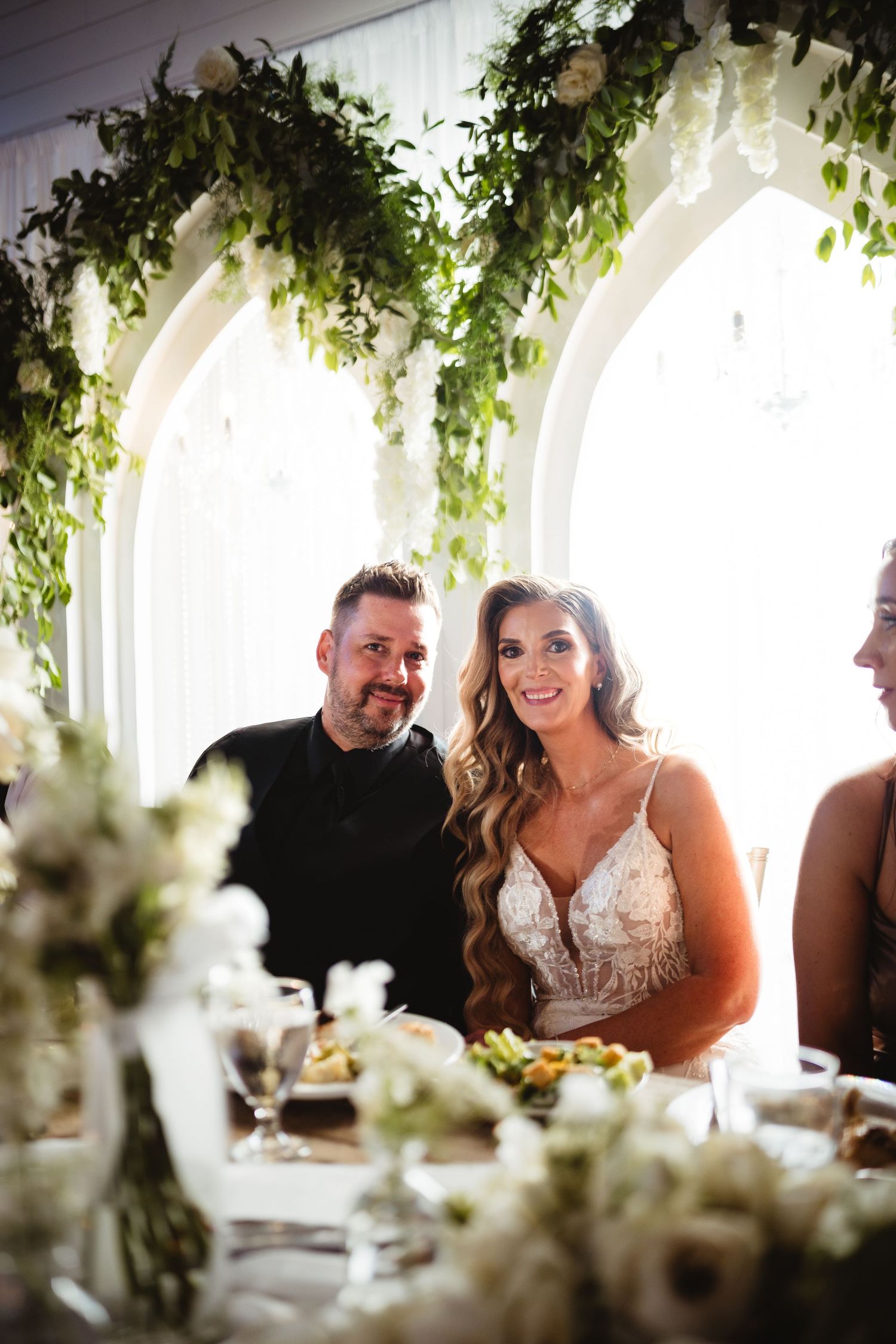 Bride and groom smiling at head table beneath lush green floral arch at their elegant wedding reception.