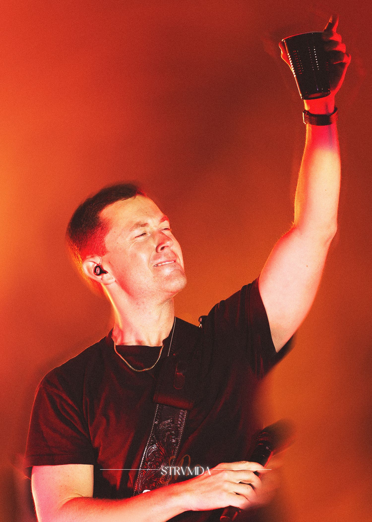 Male performer on stage raising fist under dramatic red lighting wearing black t-shirt with ear monitor.