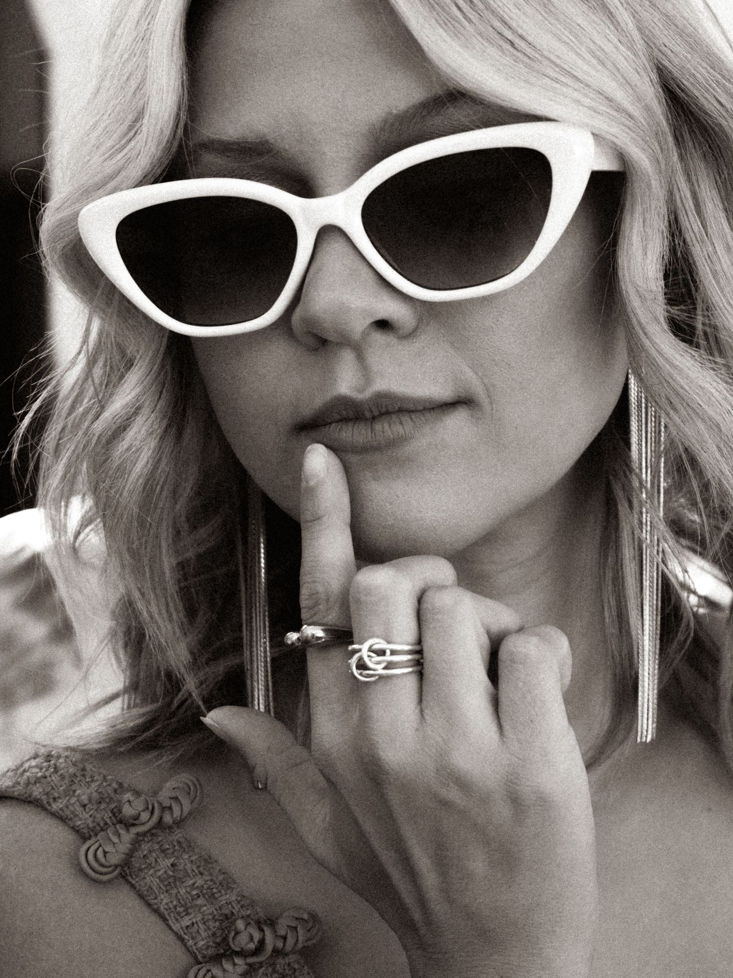 Black and white portrait of blonde woman wearing white cat-eye sunglasses and rings, posing thoughtfully.